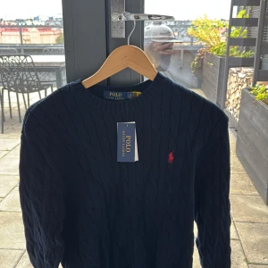 Mörkblå kabelstickad tröja Polo Ralph Lauren - Kabelstickad mörkblå tröja från Polo Ralph Lauren med klassisk rund hals och röd broderad logga på bröstet. Tröjan är långärmad och gjord i mjuk bomull, perfekt för lager på lager under kyliga dagar.