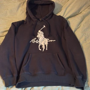Mörkblå hoodie från Ralph Lauren - Snygg mörkblå hoodie från Ralph Lauren med stort vitt Polo-logotryck på bröstet. Tröjan har huva med snörning, magficka och ribbade muddar vid ärmslut och nederkant. Perfekt för chill dagar och streetstyle. Storlek L men sitter som en M
