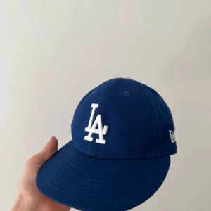 Blå LA Dodgers keps från New Era - Säljer en klassisk blå LA Dodgers keps från New Era med vit broderad LA-logga framtill och MLB-logga baktill. Kepsen har böjd skärm och är tillverkad i polyester. Perfekt för dig som gillar sportig stil och baseball.