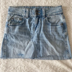 Ljusblå jeanskjol - Snygg ljusblå jeanskjol som är omsydd från ett par jeans. Kjolen har dubbla knappar framtill, bälteshällor och bakfickor med lock och knapp. Perfekt för en chill och trendig look. Kanten ser ojämn ut längst ner men är inte så när man har på sig den.