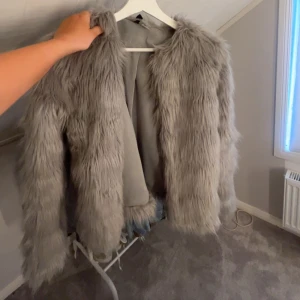 Grå fluffig pälsjacka - Säljer en supermjuk och fluffig grå pälsjacka med långärmad modell. Jackan har en oversized känsla och är perfekt för dig som vill sticka ut med en trendig look. Fodrad insida för extra komfort och värme. Passar perfekt till kalla dagar när du vill vara både varm och snygg.