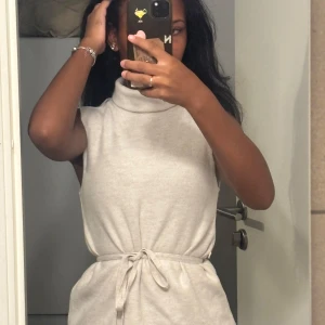 Beige ärmlös polotopp med knyt - Säljer en beige ärmlös polotopp med hög krage och knytband i midjan. Toppen har en mjuk och lite loose passform, perfekt för en chill och stilren look. Materialet känns som bomull eller en mjuk syntetmix.