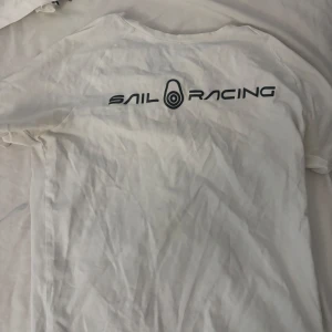 Vit t-shirt från Sail Racing - Vit t-shirt från Sail Racing med svart logotyptryck. Klassisk rund hals och korta ärmar. Perfekt för dig som gillar stilren design och sportiga märken. Materialet känns mjukt och lätt, troligtvis bomull.