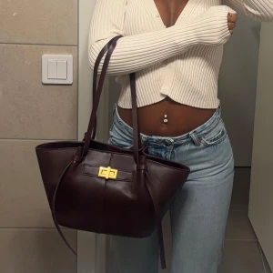 Mörkbrun skinnhandväska med guldspänne - Säljer en stilren mörkbrun handväska i skinn med snyggt guldigt spänne framtill. Väskan har långa handtag och en rymlig form, perfekt för att bära med sig allt man behöver. Enkel och klassisk design som passar till många outfits.