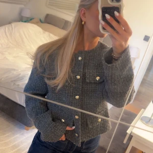 Grå cardigan jacka med guldknappar - Säljer en grå, croppad cardigan jacka med snygg struktur och guldknappar framtill. Jackan har två fickor på bröstet och raka, långa ärmar. Perfekt att slänga på över en enkel topp. Från H&M