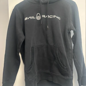 Svart hoodie från Sail Racing - Svart hoodie från Sail Racing med vit logga över bröstet. Klassisk känguruficka framtill, snörning i huvan och mjukt material. Perfekt för en avslappnad och sportig stil.