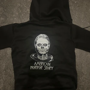 Svart American Horror Story hoodie - Svart hoodie med dragkedja och huva, tryck med American Horror Story-logga fram och stort tryck med motiv och text på ryggen. Långa ärmar och fickor framtill. Perfekt för dig som älskar serien och vill ha en cool streetstyle.