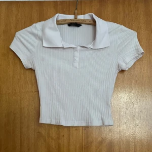 Vit ribbad croppad polotopp - Säljer en vit croppad polotopp med korta ärmar och krage. Toppen har ribbat tyg och knappar framtill vid halsen. OBS! Defekt: två små rosa fläckar framtill, se bild❤️