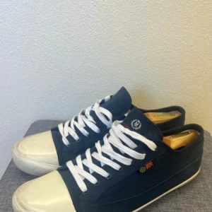 Marinblå sneakers från Henri Lloyd - Snygga marinblå sneakers från Henri Lloyd med vit tå och sula, vita snören och klassisk låg profil. Skorna har tygöverdel i bomull och en liten brittisk flagga på sidan. Perfekta för en avslappnad och stilren look. Storlek 44