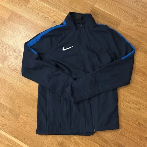 Nike Dri-Fit blå vindjacka M - Nike Dri-Fit vindjacka i marinblått med blå ränder längs axlarna och vitt Swoosh-logga på bröstet. Jackan har dragkedja framtill, hög krage och är tillverkad i lätt polyester som andas. Perfekt för träning eller när du vill ha en sportig look. Pris kan diskuteras 👍🏻👍🏻