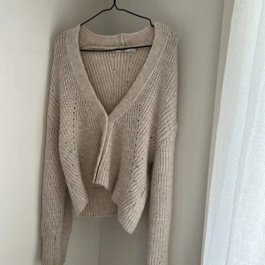 Beige stickad kofta med v-ringning - Säljer en beige stickad kofta med djup v-ringning och lång ärm. Koftan är croppad och har en loose passform, perfekt att slänga över en topp. Ribbstickade muddar och snygg struktur i materialet ger en chill vibe.
