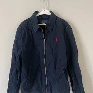 Mörkblå jacka från Polo Ralph Lauren - Klassisk mörkblå jacka från Polo Ralph Lauren med dragkedja framtill och ikonisk röd logga broderad på bröstet.Använd fåtal gånger så otroligt bra skick, nypris 1200