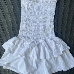 Vit kort volangklänning gina tricot - Supersöt vit kort klänning med smockad överdel och dubbla volanger nertill. Perfekt för varma dagar och ger en fräsch och somrig vibe. Axelbandslös modell som sitter snyggt och bekvämt. Endast använd en gång.