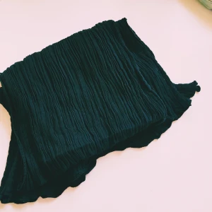 Svart plisserad halsduk - En lång och smal svart halsduk med plisserad struktur. Tunt och lätt material som ger en cool och stilren look. Perfekt att slänga över axlarna för att lyfta vilken outfit som helst.  Helt skick dock lite ljusare i färgen