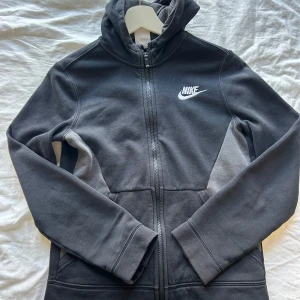 Svart zip hoodie från Nike - Svart hoodiejacka från Nike med dragkedja hela vägen och broderad vit logga på bröstet. Jackan har huva, långa ärmar och är tillverkad i mjuk bomullsmix. Inte mycket använd. Barn L men funkar som xxs