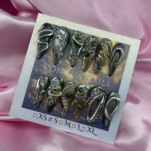 VenomCode (S);Long Stiletto  - Unika handgjorda press-on naglar i stilettoform med metallic grön, svart och silver bas. Dekorerade med 3D-detaljer i silver, kedjor och glittrande stenar i olika färger. Perfekt för dig som vill sticka ut med edgy och konstnärlig nageldesign. I storlek S: 15 mm, 11,5 mm, 12 mm, 11 mm, 9 mm 
