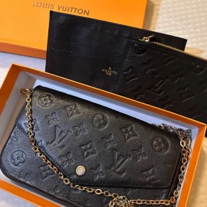 Svart axelväska i skinn - Snygg svart axelväska i präglat skinn med klassiskt monogrammönster. Väskan har en kedja och tryckknapp framtill. Kommer med matchande plånbok och innerfack, perfekt för att hålla ordning på dina grejer.