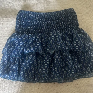 Blå mönstrad volangkjol - Supersöt trendig kortkjol med vitt diamantmönster och volanger köpt i Spanien. Kjolen har smock i midjan för en bekväm passform och är gjord i ett lätt, luftigt material. Perfekt för dig som gillar en lekfull och trendig stil. Det är även inbyggda shorts vilket är jätteskönt. Bara använd en gång 