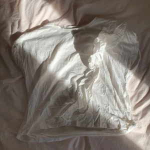 Vit t-shirt med volangdetalj - Säljer en vit t-shirt i bomull med korta ärmar och en snygg volangdetalj längs ena axeln. T-shirten har en klassisk rund halsringning och är enkel men ändå unik tack vare volangen. Perfekt för dig som gillar stilrena plagg med en twist.