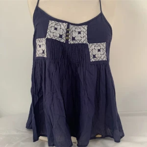 Mörkblå topp med spetsdetaljer - Snygg mörkblå ärmlös topp med vita spetsdetaljer och knappar framtill. Den har smala axelband och en luftig passform, perfekt för varma dagar. 