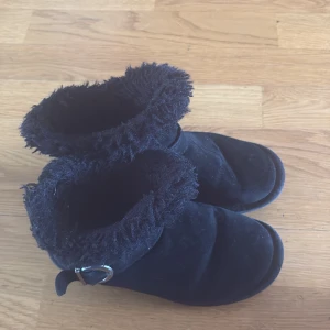 Svarta boots med fuskpäls och spänne - Mysiga svarta boots med fluffig fuskpäls runt skaftet och dekorativt spänne på sidan. Skorna har rund tå och platt sula, perfekta för kalla dagar. Utsidan är i mockaliknande material och insidan är fodrad för extra värme.