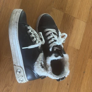 Svarta sneakers med päls från Guess - Coola svarta sneakers från Guess med vit fuskpäls runt öppningen och vita snören. Sulan är dekorerad med stora glittriga stenar och Guess-logga på sidan och baksidan. Skorna har en chunky look och är perfekta för dig som vill sticka ut.