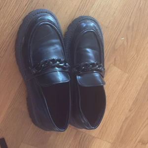 Svarta chunky loafers med kedjedetalj - Snygga svarta loafers med chunky sula och cool kedjedetalj över foten. Skorna har rund tå och är tillverkade i glansigt syntetmaterial som ger en modern look. Perfekta för dig som vill sticka ut med din stil.