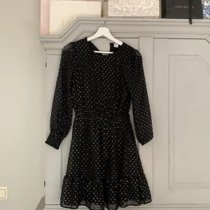 Svart prickig klänning från Lindex - Supersöt svart kortklänning från Lindex med glittriga prickar, långärmad och med resår i midjan. Volang nertill ger extra flow och ärmarna är lätt puffade. Perfekt för dig som gillar lite extra glitter i vardagen!