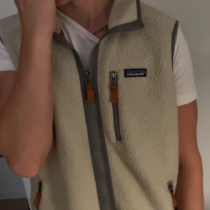 Beige fleeceväst från Patagonia L - Säljer en beige fleeceväst från Patagonia i storlek L. Västen har grå detaljer, hög krage och tre fickor med dragkedja och bruna läderdetaljer. Perfekt att slänga över en hoodie eller t-shirt för extra värme. Klassisk Patagonia-logga på bröstet.