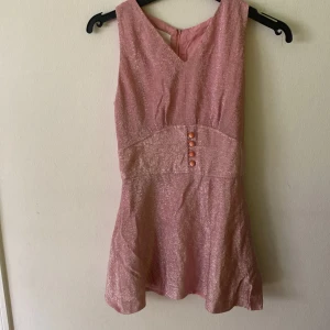 Rosa glitter blus. Elson of Denmark - Supersöt och festlig rosa blus från Elson of Denmark.  Troligtvis 60-tal  med glittrigt tyg och fyra dekorativa knappar i midjan. Klänningen är ärmlös, har v-ringning och markerad midja. Perfekt för dig som vill sticka ut med en unik vintage-look!  Är egentligen en 40 men väldigt liten i storlek så skulle snarare räknas som xs/s.