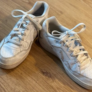 Vita Nike sneakers i skinn - Klassiska vita Nike sneakers i skinn med perforerade detaljer och snörning. Snygg låg modell med rund tå och diskret Nike-logga på sidan. Perfekta för en clean och sportig look.
