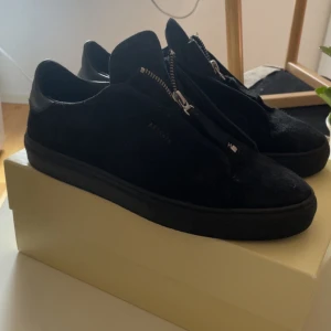Arigato skor - Stilrena svarta sneakers från Axel Arigato i mjuk mocka med coola dragkedjor framtill istället för klassisk snörning. Skorna har rund tå, platt sula och diskret logga på sidan. Perfekta för dig som gillar minimalistisk och modern design.