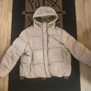 Beige pufferjacka med huva - Säljer en beige pufferjacka med huva och dragkedja.  Passformen är större i storleken och jackan är lätt att bära.