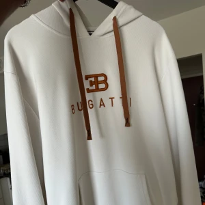 Vit hoodie från Bugatti med bruna detaljer - Snygg vit hoodie från Bugatti med bruna snören och broderad logga på bröstet. Klassisk känguruficka framtill och huva. Perfekt för dig som gillar stilrena och exklusiva streetwear-plagg.