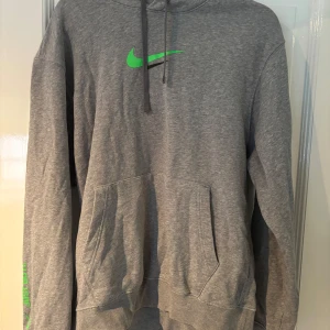Grå Nike hoodie med gröna detaljer - Snygg grå hoodie från Nike med grönt och svart tryck på bröstet och ryggen. På ärmen står det 'Just Do It.' i grönt. Tröjan har huva med snörning och en stor magficka. Perfekt för dig som gillar sportig och avslappnad stil.