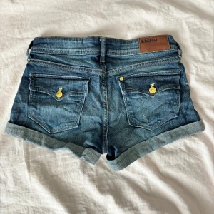 Blå jeansshorts från H&M - Snygga blå jeansshorts från &Denim med uppvikta benslut och klassiska guldiga knappar. Shortsen har fem fickor och bälteshällor, perfekt för en chill sommarlook. Tidlös denim med coola detaljer och skön passform.