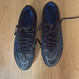 Nike fotbollsskor air soom PRO  - Säljer ett par svarta Nike fotbollsskor med blå och vita detaljer. Skorna har snörning, platt sula med dobbar och en stickad strumpkrage för extra stöd. Ovandelen är i syntetmaterial och sulan har blå inslag. Perfekta för dig som vill ha stil på planen.