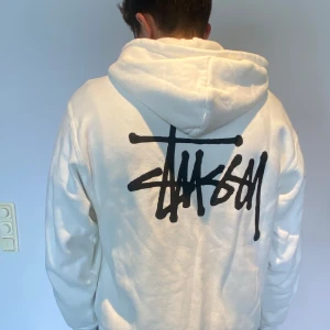 Vit hoodie med dragkedja från Stüssy - Säljer en vit hoodie från Stüssy med dragkedja framtill och stor svart logga på ryggen. Ordinarie pris är 1827kr från stussy.com. Tröjan är i mycket bra skick och har blivit använd 2 gång. Tröjan har huva med snörning, två fickor fram och mindre logga på bröstet. Perfekt för dig som gillar streetwear och vill ha en clean look. 