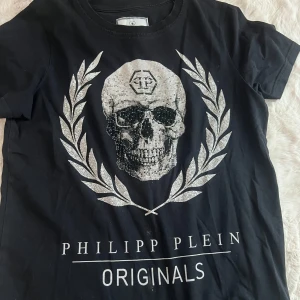 Svart t-shirt Philipp Plein Junior - Svart t-shirt från Philipp Plein Junior med stor glittrig döskalle och silverfärgade lagerblad på bröstet. 