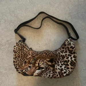 Axelväska med leopardmönster - Cool axelväska med leopardmönster och ett stort tryck av en gepard på framsidan. Väskan har justerbar svart axelrem och dragkedja upptill samt en extra ficka med dragkedja på framsidan. Perfekt för dig som vill sticka ut med din stil.