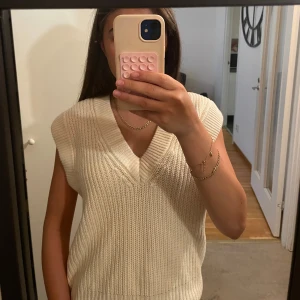 Vit stickad väst med v-ringning - Säljer en krämvit stickad väst med bred v-ringning och ribbad struktur. Västen är ärmlös och har en avslappnad passform som är perfekt att styla över en skjorta eller t-shirt. Enkel och stilren design som funkar året runt.