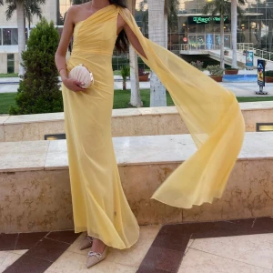Gul långklänning med oneshoulder - Superfin gul långklänning med oneshoulder och svepande cape-detalj. Klänningen har draperad överdel och markerad midja, vilket ger en elegant och flowy siluett. Perfekt för bal eller fest där du vill sticka ut med en unik look.