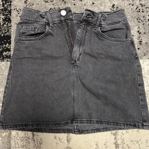 Grå jeanskjol från H&M Denim - Snygg mörkgrå jeanskjol från H&M Denim med klassisk femficksdesign, bälteshällor och dragkedja framtill. Kjolen är i kort modell och har justerbar midja på insidan för perfekt passform. Perfekt till sneakers eller boots.