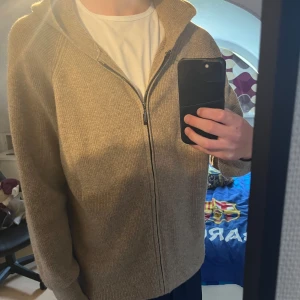 Beige stickad hoodie med dragkedja - Klockren beige cashmere hoodie med hel dragkedja framtill och huva. Tröjan har en enkel och stilren design med ribbade detaljer längs ärmar och axlar. Perfekt nu inför hösten. Nästintill oanvänd. Priset ej hugget i sten. Hör av er vid minsta lilla fundering.