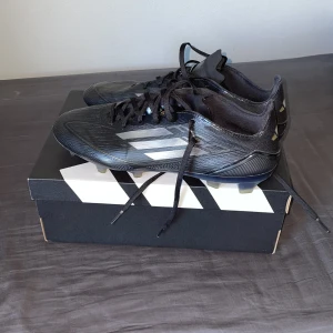 Svarta adidas fotbollsskor med dobbar - Snygga svarta adidas fotbollsskor med klassiska tre ränder i silver på sidan. Skorna har snörning och platt sula med fasta dobbar för bra grepp på planen. Tillverkade i syntetmaterial och designade för att vara lätta och snabba på fotbollsplanen. Det är inte några elite skor det är pro. Skorna är oanvända, har endast tagit på mig dem för att kolla ifall dem passade eller inte. Dem var för stora för mig. Går också med på att göra byten på fotbollsskor och kan diskutera priset.