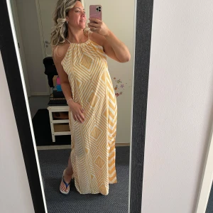 Gulvit långklänning med grafiskt mönster - Säljer en flowy långklänning i vitt och gult med grafiskt, randigt mönster. Klänningen har halterneck och är ärmlös, perfekt för varma dagar. Lätt och luftig passform som ger en chill vibe. Superfin till sandaler eller sneakers!