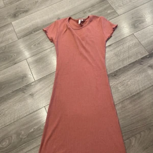 Rosa ribbad långklänning - En enkel och snygg långklänning i ribbad rosa bomull. Klänningen har rund halsringning och korta ärmar, perfekt för en avslappnad och trendig look. Passformen är normal och materialet känns mjukt mot huden.