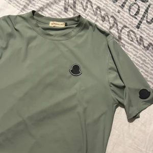 Grön Moncler t-shirt med logga - Snygg grön t-shirt från Moncler med svart logopatch på bröstet och ärmen. T-shirten har en klassisk passform, rund hals och är tillverkad i ett mjukt, ribbat material som känns riktigt skönt mot huden.