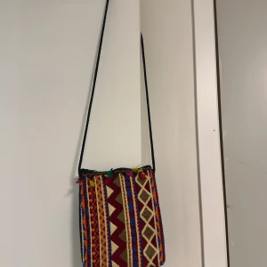 Färgglad axelväska med mönster - Unik axelväska med färgstarkt geometriskt mönster i rött, blått, gult och beige. Väskan har lång svart axelrem och är tillverkad i ett vävt bomullstyg. Perfekt för dig som vill sticka ut med en bohemisk och kreativ stil.