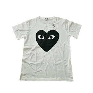 Vit Comme des Garçons Play t-shirt - Helt ny med tag, Vit t-shirt från Comme des Garçons Play med stort svart hjärta och ögon-tryck på bröstet. Klassisk rund halsringning och korta ärmar. Tillverkad i mjuk bomull, perfekt för en clean och cool look. 